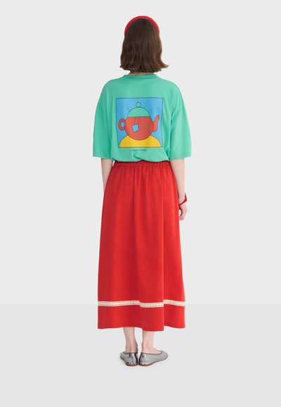 Femme de dos portant une chemise bleu sarcelle avec un graphique de théière colorée, une jupe midi rouge avec une bordure blanche, un bandeau rouge et des ballerines argentées.