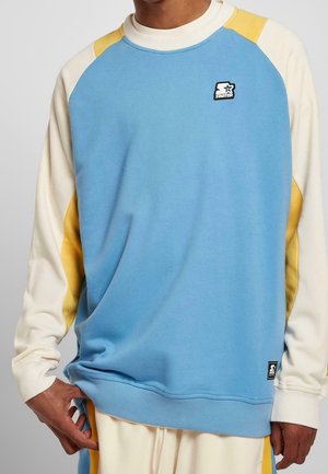 Hombre vistiendo un sudadera de bloques de color con la parte delantera azul, mangas color crema y acentos amarillos, sosteniendo el dobladillo de la sudadera cerca de unos pantalones a juego.