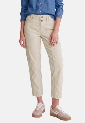 Femme portant un pantalon beige court à double boutons, une chemise bleue à manches longues et des baskets lacées rose clair sur fond blanc.