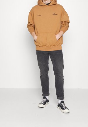 Mustard-colored hoodie with a front pocket, black text on the chest, paired with dark jeans and black sneakers with white soles.