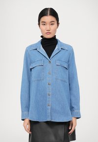 Helleblaue Jeanshemd mit langen Ärmeln, Knopfschluss, zwei Brusttaschen und einem Kragen mit Spitze. Wird über einem schwarzen Rollkragenpullover getragen.