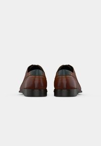 Un paio di scarpe di pelle marroni con tacco nero viste da dietro su uno sfondo bianco.