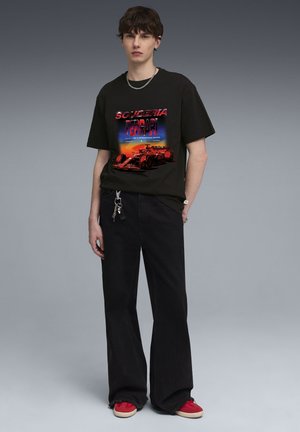 Schwarzes Baumwoll-T-Shirt mit einem bunten Ferrari-Motiv. Kombiniert mit schwarzen, weit geschnittenen Hosen und roten Sneakers mit beigen Sohlen.