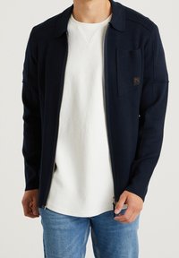 Marinblå zip-jacka med krage, en bröstficka och textilstruktur; bärs över en vit crew-neck tröja och blå denimjeans.