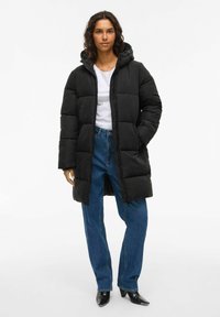 Sieviete valkā melnu pufīgu mēteli ar kapuci virs baltas krekla, zilu džinsu un melnām šaurām zābakiem, stāvot pret vienkāršu fona.