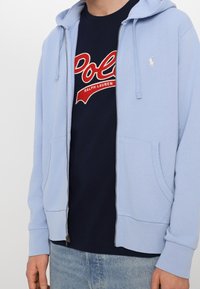 Ljusblå zip-up hoodie med kängurufickor, ribbade ärmslut och nederkant. Bärs över en marinblå t-shirt med röd broderad text.