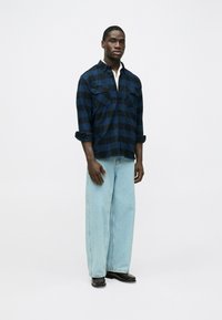 Camicia di flanella a quadri blu e nera con due tasche sul petto, maniche arrotolate, abbinata a jeans a gamba larga azzurri e stivali scuri.