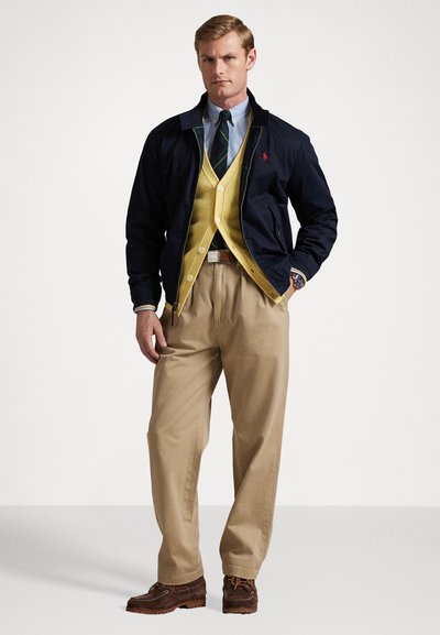 Marineblaues Jacket mit grünen Akzenten, geleste Weste über blauem Hemd, beige Hosen, braune Schuhe. Klassisches Design mit Knopf- und Taschendetails.