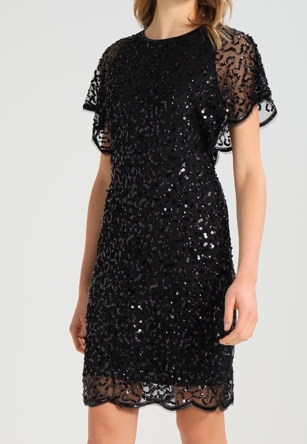 Femme portant une robe noire à sequins avec des manches courtes transparentes et un ourlet festonné, debout devant un fond uni.