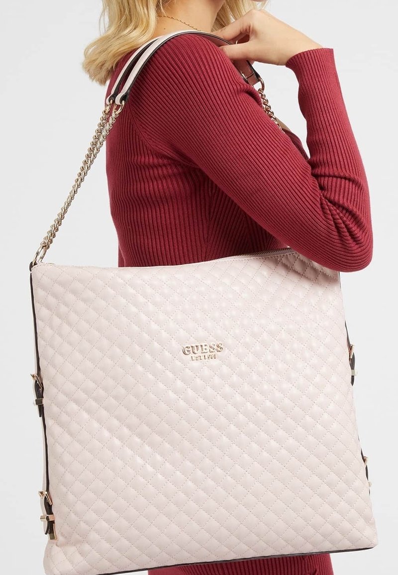 Kobieta w czerwonym prążkowanym swetrze, niosąca dużą pikowaną torebkę Guess w jasnoróżowym kolorze z złotym łańcuchem i detalami logo.