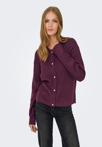 Violette Strickjacke mit langen Ärmeln, Knopfverschluss mit kleinen weißen Knöpfen und leicht gekürztem Saum. Dazu dunkelblaue Jeans getragen.