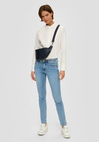 Zwarte leren schoudertas met ritssluiting, gedragen over een witte blouse, in combinatie met lichtblauwe skinny jeans en witte sneakers.