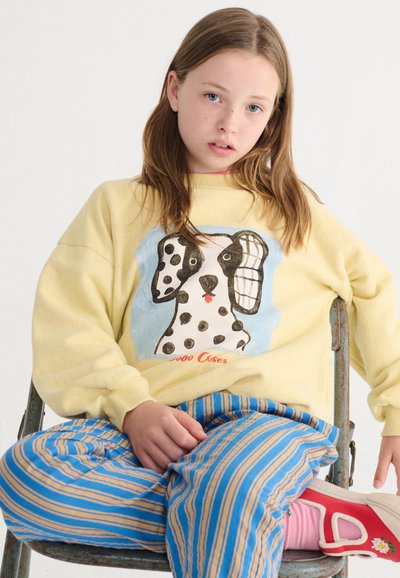 Mladá osoba s dlhými vlasmi sediaca, oblečená v žltej mikine s grafikou škvrnitého psa, modrými a žltými pruhovanými nohavicami a červenými topánkami s ružovými ponožkami.