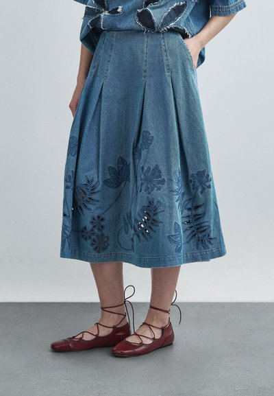 Jupe en denim bleu clair à plis, ornée de motifs floraux découpés. Associée à des chaussures plates rouges à lacets. Texture mate.