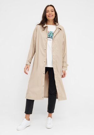 Beige langem Regenmantel aus glattem Material mit Knopfleiste, übergroßem Kragen und hochrollbaren Ärmeln, kombiniert mit einem weißen Grafik-T-Shirt und dunklen Jeans.