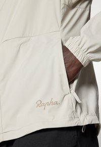 Jasno-beżowa kurtka wykonana z gładkiego materiału. Posiada elastyczne mankiety, boczną kieszeń oraz subtelne logo "Rapha" w jaśniejszym odcieniu.
