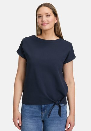 Marineblaues kurzärmeliges Oberteil mit rundem Halsausschnitt, strukturiertem Stoff und einer gebundenen Detail am Taillenbereich, kombiniert mit hellblauen Jeans.