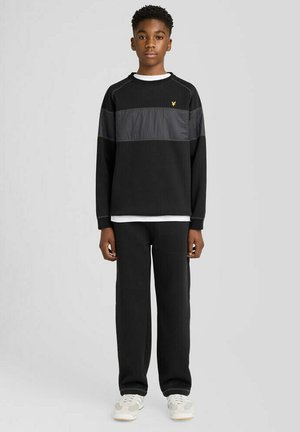 Tienerjongen staat, draagt een zwart shirt met lange mouwen met een grijze streep en zwarte broek, witte sneakers, op een effen lichtgrijze achtergrond.
