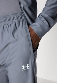 Sive trenirke od mrežaste tkanine, s elastikom u struku i bočnim džepovima. Na donjem dijelu nogavice se nalazi bijeli logo Under Armour.