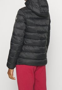 Person som bär en svart vadderad jacka med huva och logotyp på ärmen, i kombination med röda byxor, sedd bakifrån mot en enfärgad bakgrund.