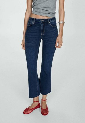 Person iført mørkeblå korte jeans, grå ærmeløs top, røde netflade sko med ankelremme og sølv manchetarmbånd på venstre arm.