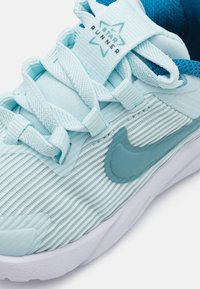 Lys blå idrettssko med teksturerte striper, med en teal aksent og en fremtredende Nike-logo på siden. Snøringene er stramt festet.