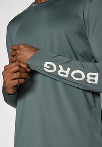 Dlhý rukáv, teal atletické tričko s okrúhlym výstrihom a bielym perforovaným textom "BORGC" na rukáve. Hladká textúra, ľahký materiál.