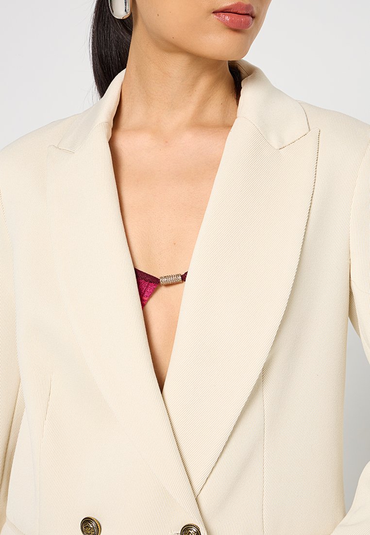 Blazer a coste crema con revers appuntiti e dettagli in bottone dorato, che mette in mostra un profondo scollo a V su un reggiseno rosso con dettagli metallici.