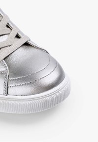 Zapatilla metálica plata con superficie texturizada, punta redondeada y cordones beige. La suela de goma blanca presenta un patrón de diamante para mayor tracción.
