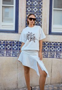 Kvinde iført oversized solbriller, hvid t-shirt med rosenprint, asymmetrisk hvid nederdel med slids, stående ved en blå mønstret flisevæg.