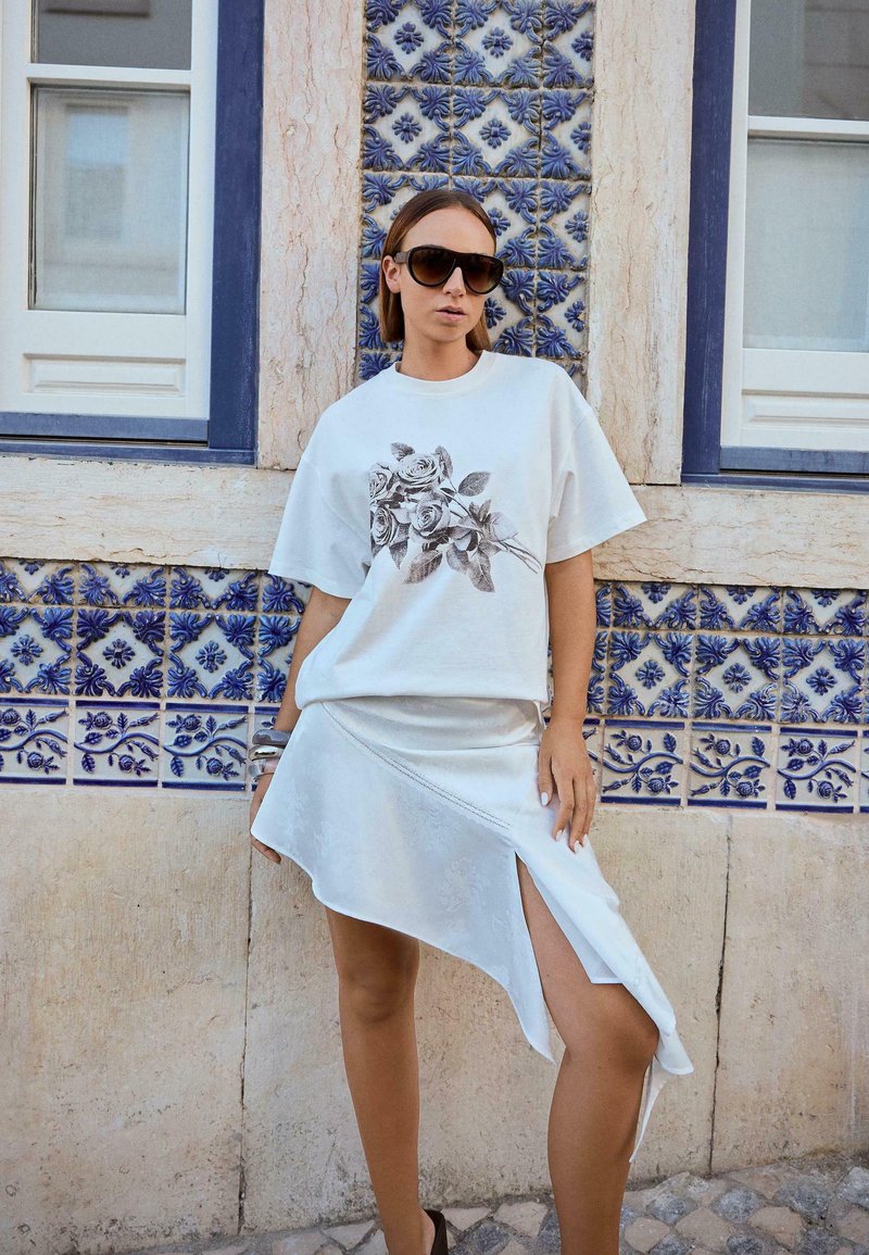 Kvinde iført oversized solbriller, hvid t-shirt med rosenprint, asymmetrisk hvid nederdel med slids, stående ved en blå mønstret flisevæg.