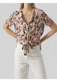 Donna che indossa una blusa a maniche corte con bottoni, stampata con fiori marroni, neri e rosa, annodata in vita, abbinata a pantaloni bianchi a vita alta.