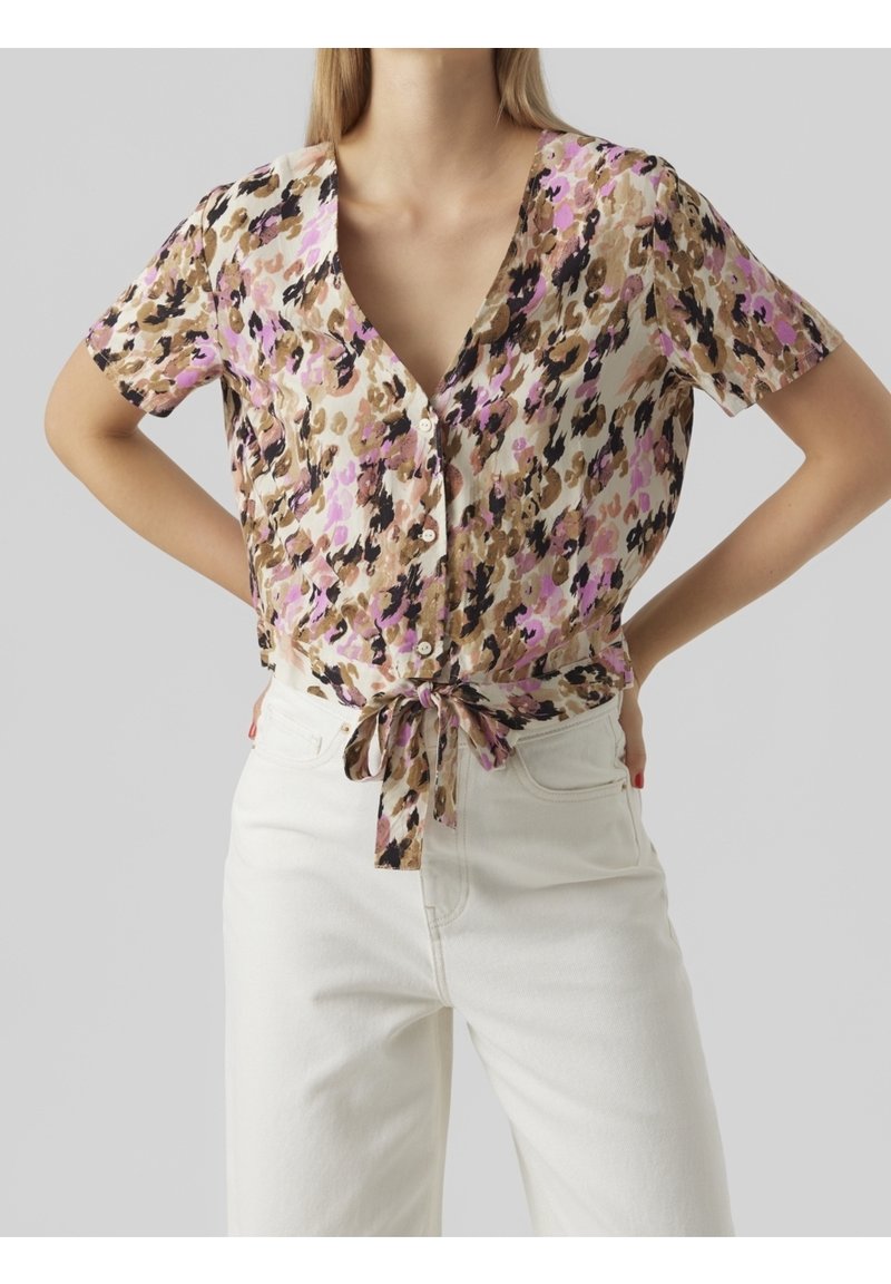 Donna che indossa una blusa a maniche corte con bottoni, stampata con fiori marroni, neri e rosa, annodata in vita, abbinata a pantaloni bianchi a vita alta.