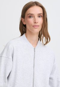Veste bomber zippée gris clair avec un col rond, des épaules tombantes et une texture douce ; elle présente une fermeture éclair lisse et des détails de couture minimalistes.