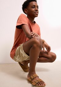 Bruna mockasiner med dubbla spännen, beiga broderade shorts och en korallfärgad t-shirt. Personen sitter hukad mot en neutral bakgrund.