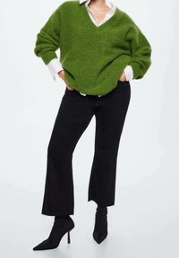 Pull vert surdimensionné et duveteux avec col en V, porté par-dessus une chemise blanche. Associé à un jean noir évasé et des bottines noires pointues.