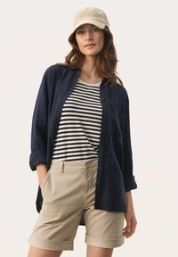 Camicia in lino blu sopra una maglietta a righe bianche e nere, abbinata a pantaloni corti color khaki chiaro. Presenta maniche arrotolate e tasche. Cappellino beige.