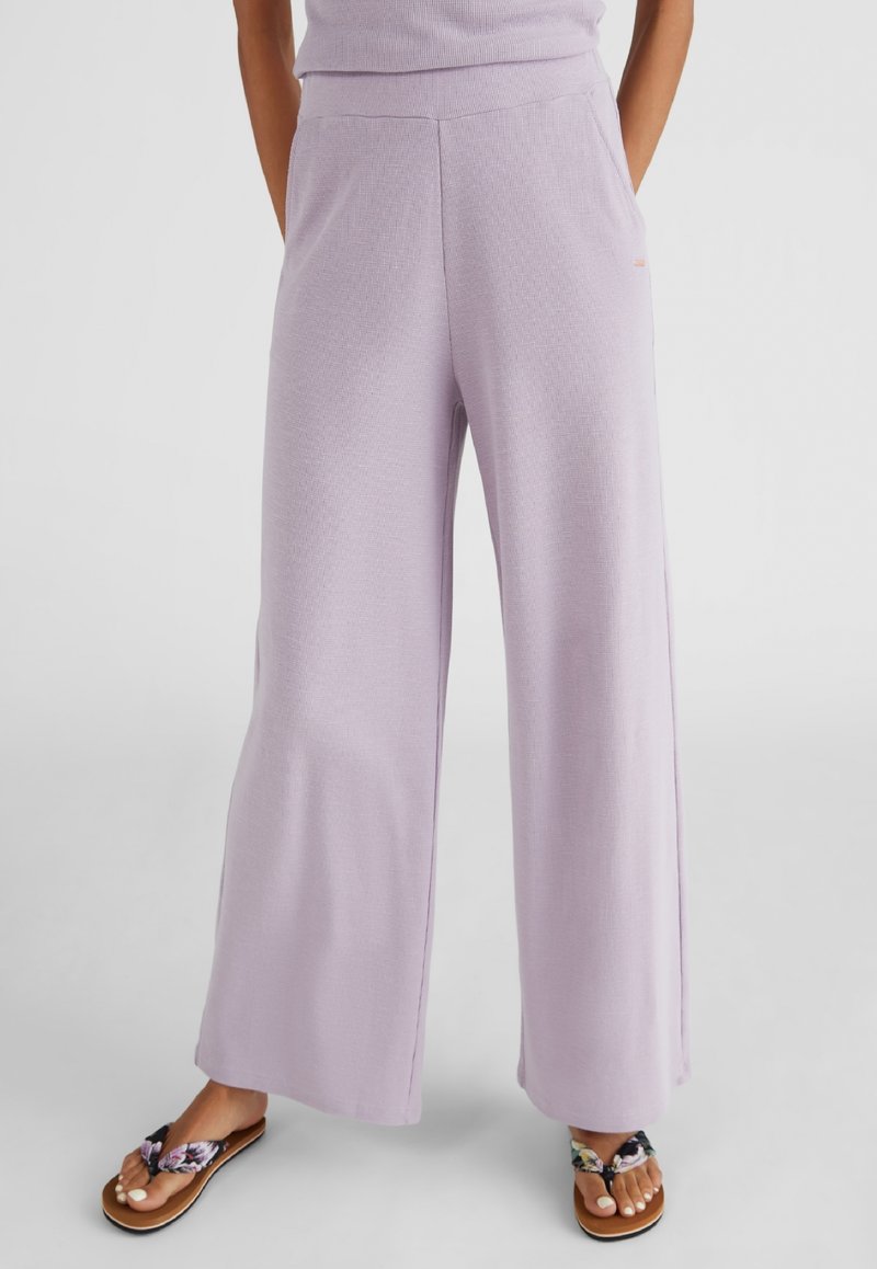 Pantalons larges en lavande clair en tissu texturé, avec une taille élastique et des poches latérales. Associés à des tongs fleuries.