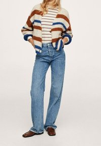 Sweter w kolorze kremowym ze brązowymi i niebieskimi paskami, zestawiony z paskowaną koszulą i szerokimi niebieskimi dżinsami. Brązowe, teksturowane buty dopełniają stylizację.