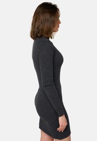 Femme avec des cheveux bruns jusqu'aux épaules, portant une robe en tricot ajustée à manches longues de couleur gris foncé, se tenant de profil contre un fond uni.