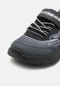 Svart atletisk sko med en texturerad gummisula, hexagonalt mönster och silverdetaljer. Har elastiska snören och ett kardborreband.