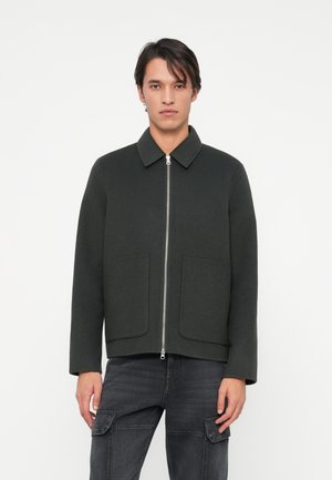 Veste zippée verte foncée avec un col, deux poches avant et une silhouette décontractée. Fabriquée dans un tissu texturé semblable à de la laine.