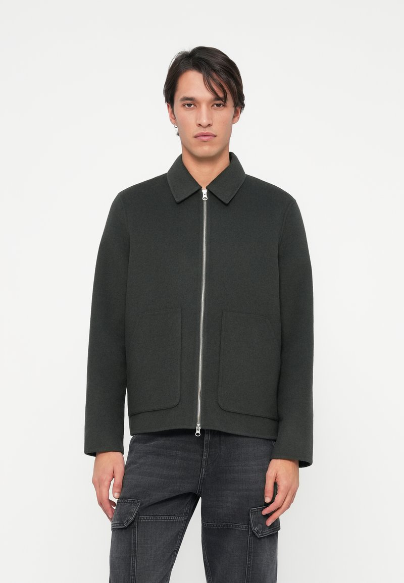 Dunkelgrønn, jakke med glidelås og krage, to frontlommer og en avslappet silhuett. Laget av et ullaktig tekstil.