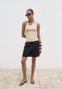 Top a righe beige abbinato a pantaloni corti neri su misura. Indossato con occhiali da sole neri e una collana floreale, con uno sfondo neutro.