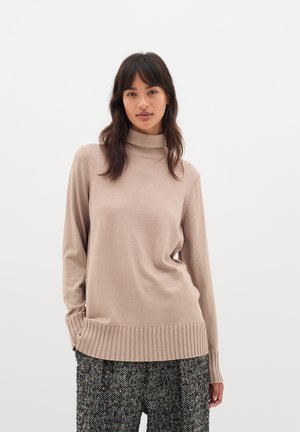 Beige turtleneck sweater with long sleeves and ribbed hem, made of a soft material, worn over patterned black and white trousers.