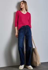 Różowa bluzka z długim rękawem i dekoltem w kształcie V, połączona z ciemnoniebieskimi spodniami w szerokim kroju oraz białymi trampkami. Niesie beżową torbę teksturowaną.