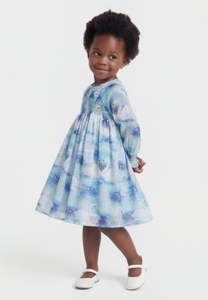 Ung pige iført en blå og lilla svanemønstret kjole med lange ærmer og hvide sko, stående med hænderne bag ryggen og smilende.