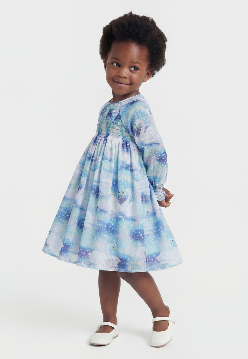 Fată tânără care poartă o rochie albastră și violetă cu model de lebădă, mâneci lungi și pantofi albi, stând cu mâinile la spate și zâmbind.