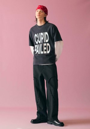 Jeune personne debout devant un fond rose, portant un bandana rouge, un t-shirt noir oversize avec le texte "CUPID FAILED", un pantalon noir et des chaussures noires.