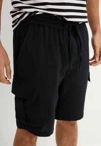 Pantaloni cargo neri realizzati in tessuto strutturato. Presentano una vita elasticizzata, un cordoncino e tasche laterali con una vestibilità comoda.